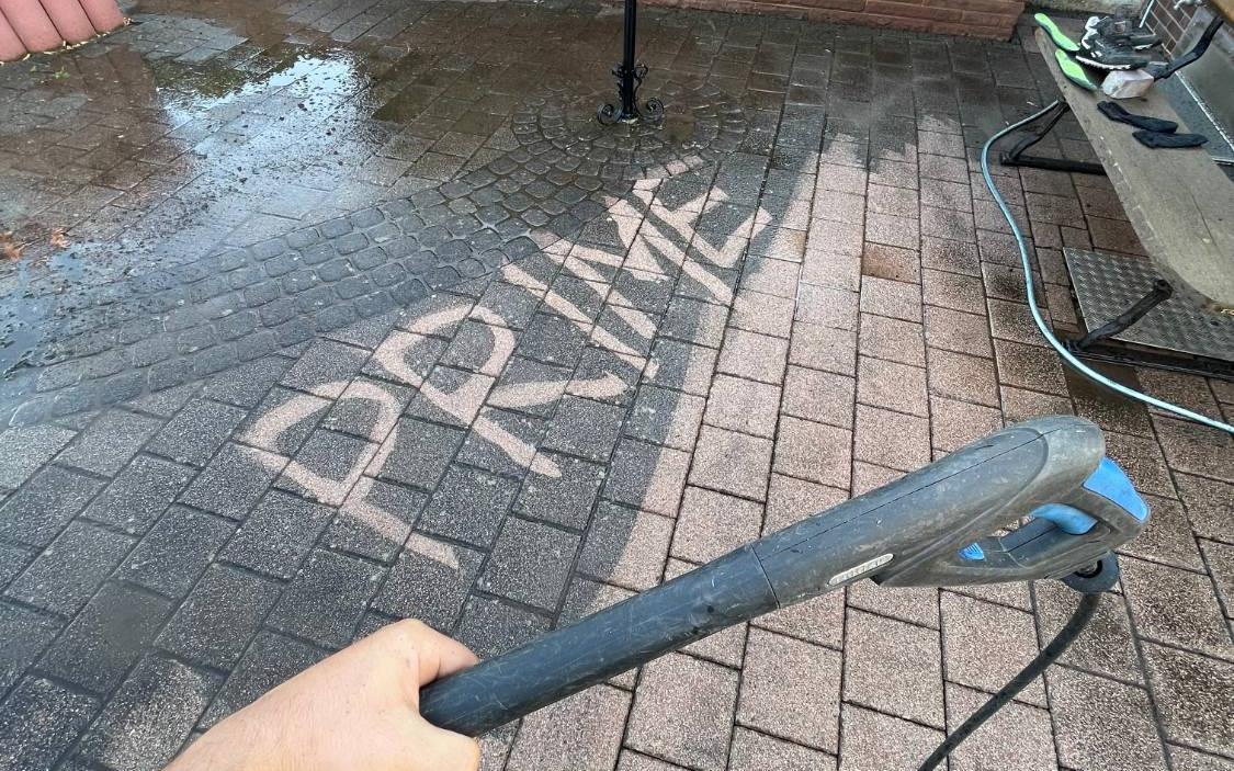 Hand mit Hochdruck-Pistole über einem Hof: Auf dem grauen Pflaster steht hell herausgereinigt der Schriftzug PRIME